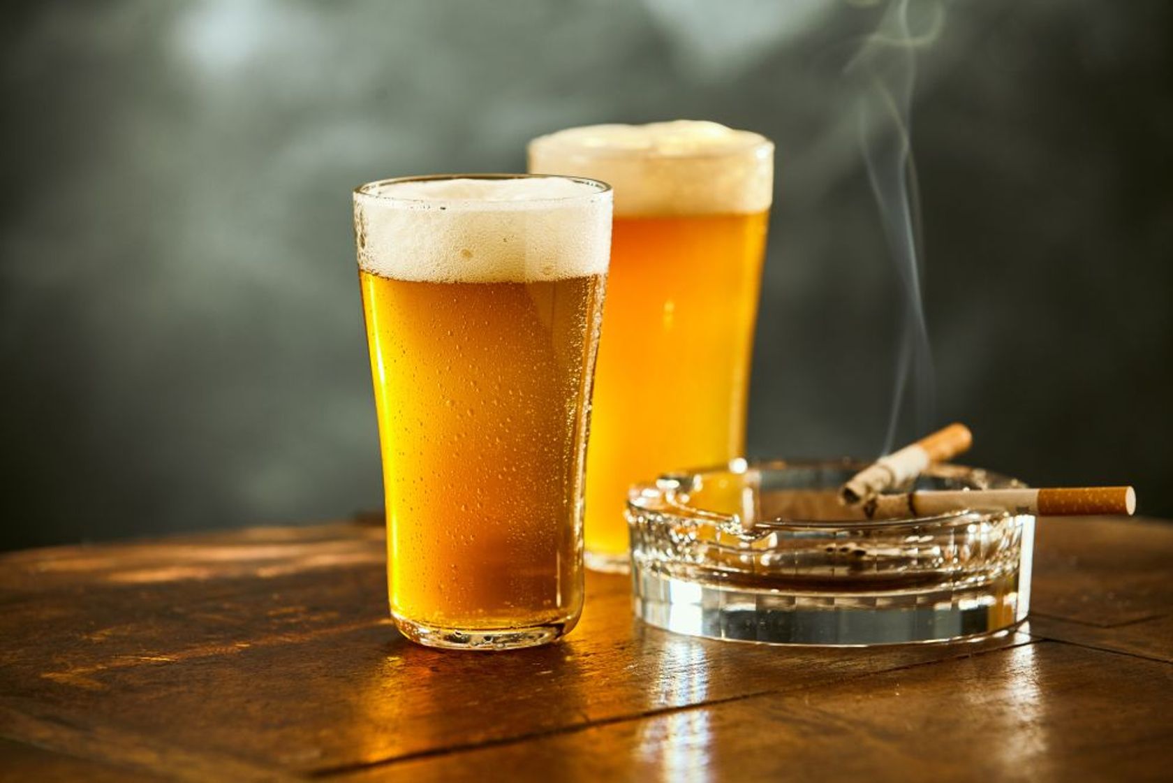 Zwei volle Biergläser und zwei brennende Zigaretten im Aschenbecher auf einem Tisch.