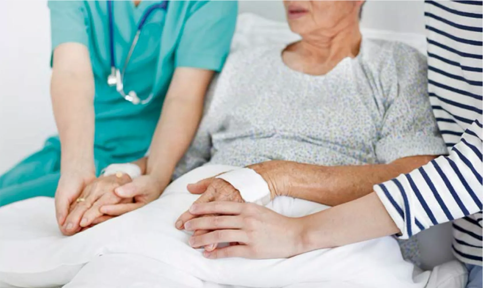 Ärztin und Angehörige sitzen am Bettrand einer kranken Frau und halten ihre Hände.