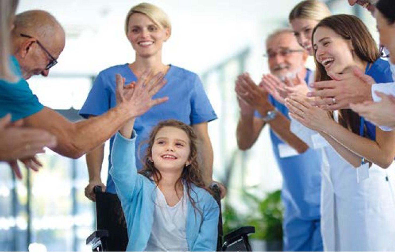 Ein blasses Mädchen sitzt im Rollstuhl und wird umringt von medizinischem Personal in blauer Arbeitskleidung, die lachen und ihr applaudieren.