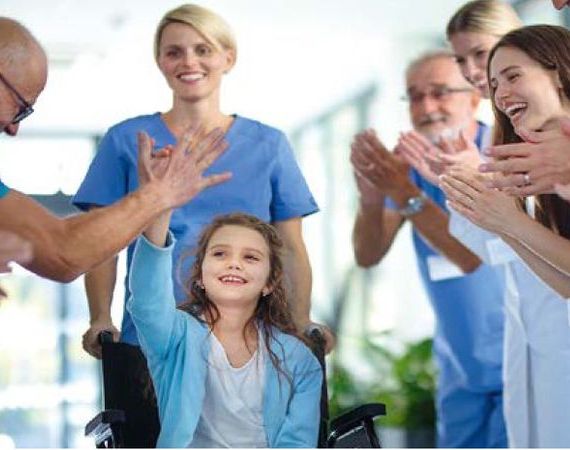 Ein blasses Mädchen sitzt im Rollstuhl und wird umringt von medizinischem Personal in blauer Arbeitskleidung, die lachen und ihr applaudieren.