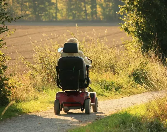 Eine Seniorin fährt mit einem Elektrorollstuhl auf einem Feldweg.